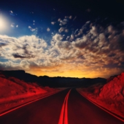 A desert road, lit with red in the foreground, with the glowing lights of Las Vegas in the background