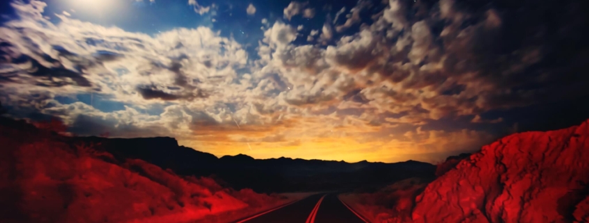 A desert road, lit with red in the foreground, with the glowing lights of Las Vegas in the background
